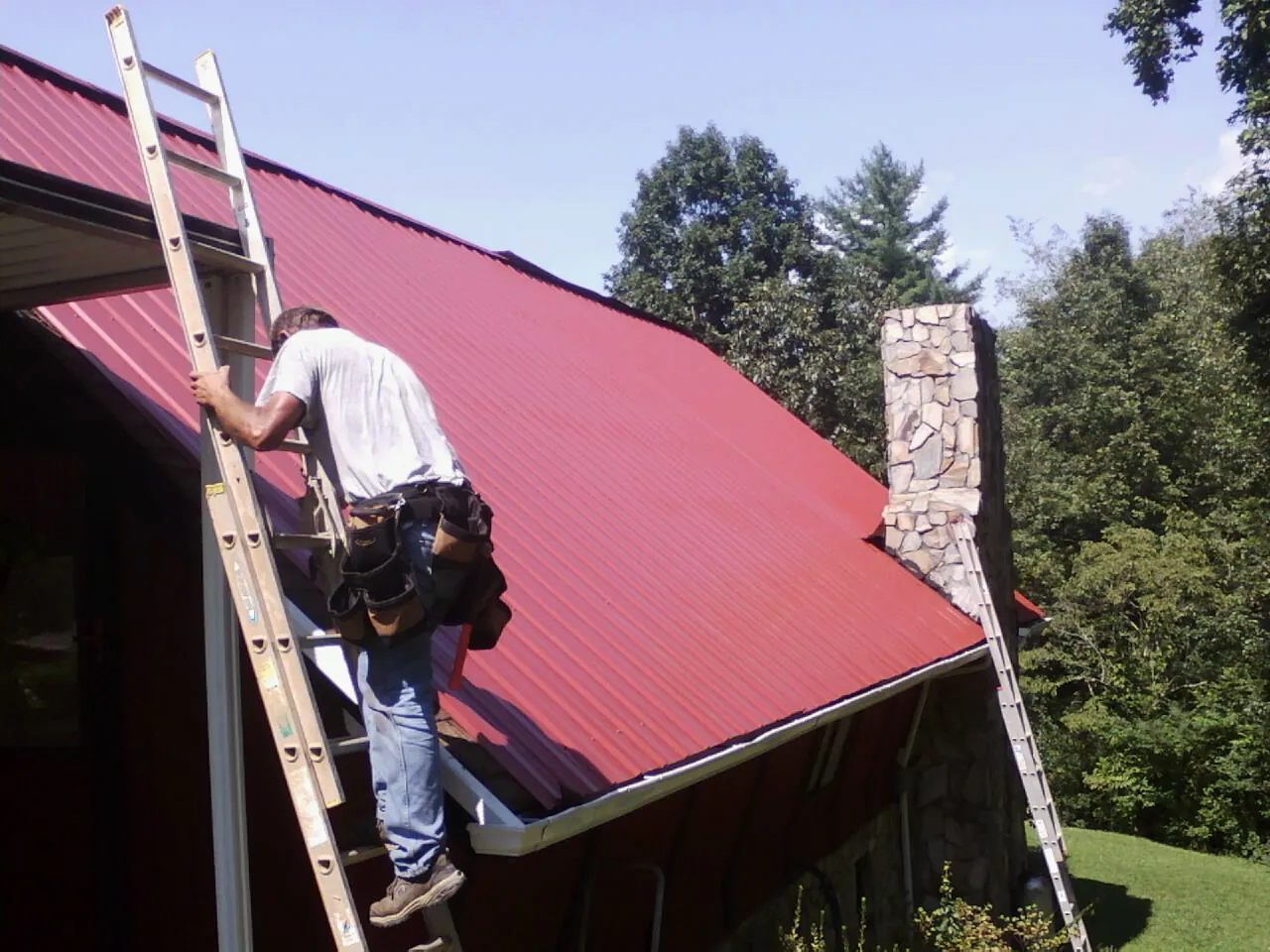 Roofer on a ladder beside a red metal roof and stone chimney — Soffit & Fascia Repair by Roofing Brothers in Fentress